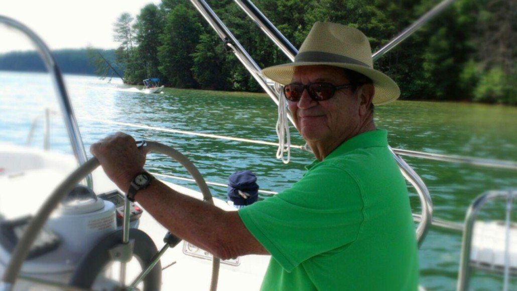 Man in a straw hat and sunglasses steers a sailboat on a lake, smiling.