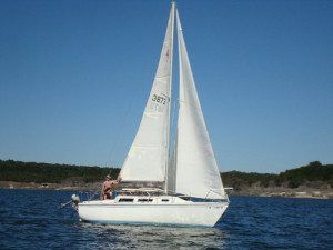 White sailboat sailing on blue water under a clear sky. A person stands at the helm.