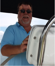 Man with sunglasses steering a boat, wearing a blue shirt.