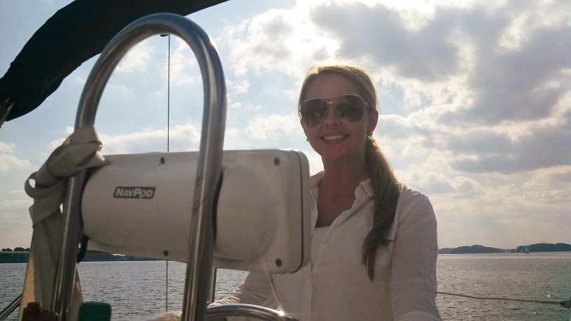 Woman wearing sunglasses smiles while sailing a boat on a sunny day.