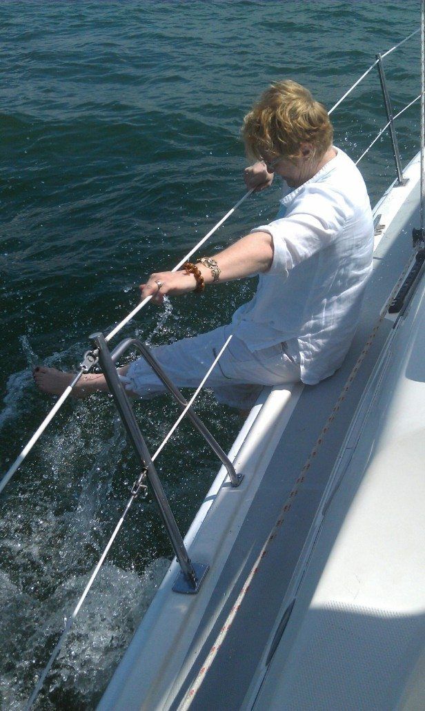 Person sitting on side of a sailboat, holding onto a railing, with feet dangling towards the water.