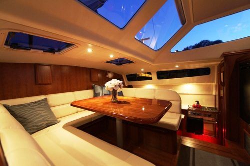 Interior of a sailboat cabin, showcasing a wooden table, cream-colored seating, and a view of the sky through the windows.