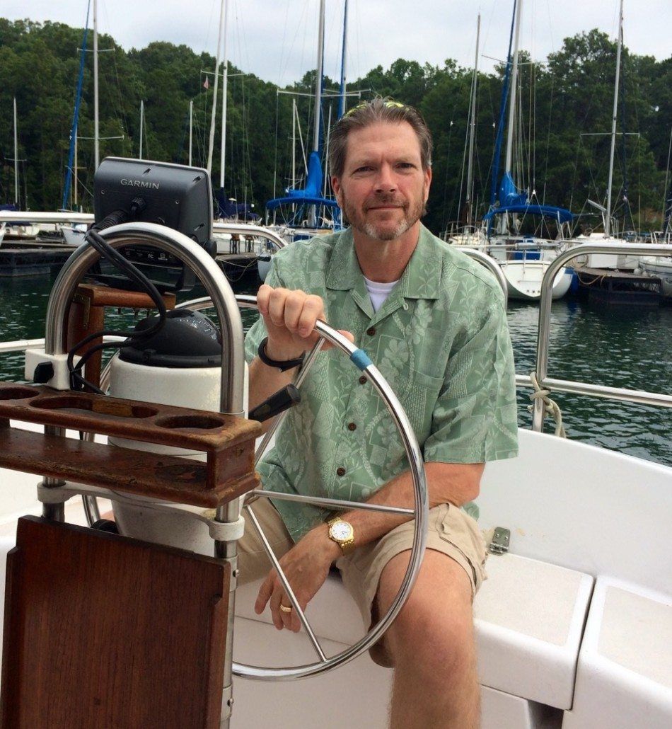 Man steering a sailboat, wearing a green shirt and khaki shorts. Setting: marina with sailboats.