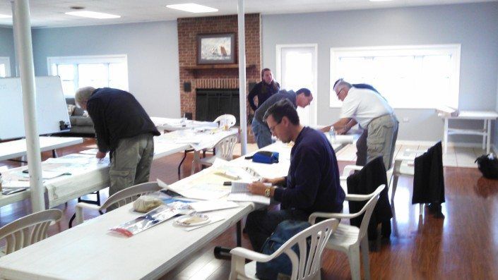 Men at tables in a room, focused on papers, possibly a meeting or workshop. White tables, wood floors, fireplace.
