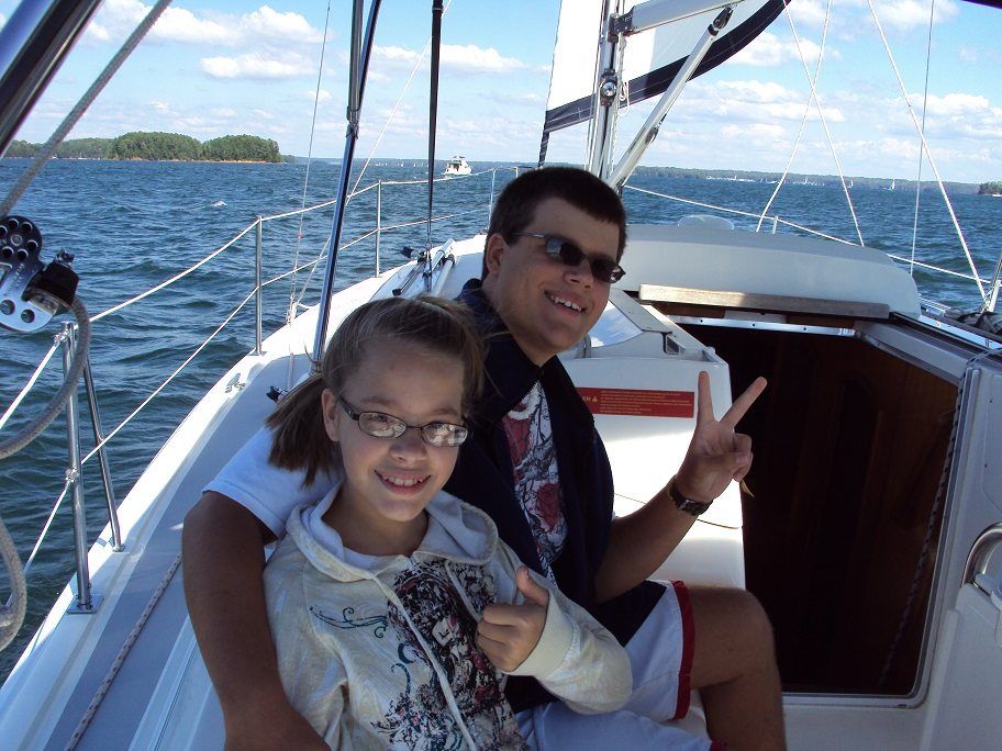 Father and child sailing on a boat, smiling at the camera. Sunny day on water, child gives thumbs up, father peace sign.