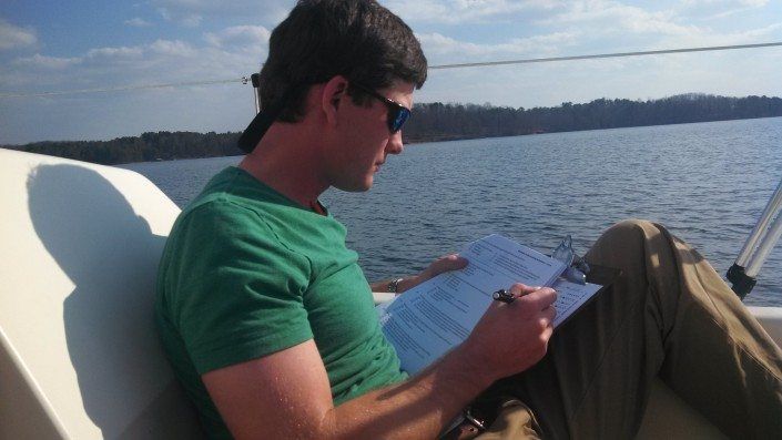 Man in sunglasses and green shirt writing on a paper on a boat, lake in background.