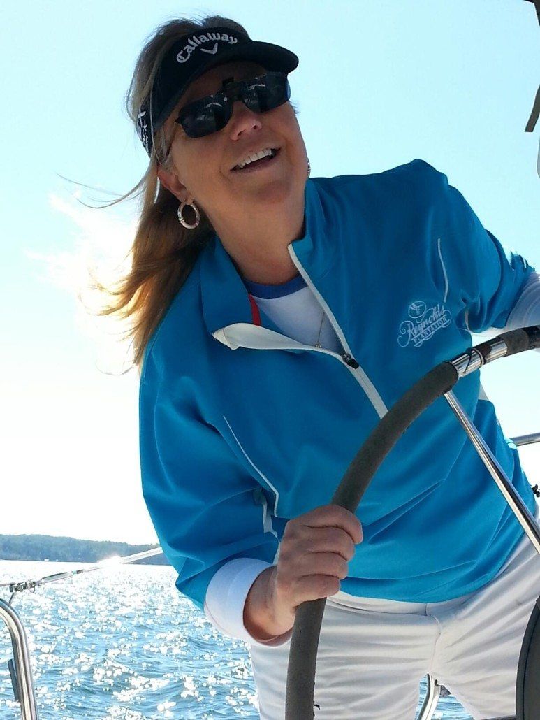 Woman wearing a visor, sunglasses, and blue jacket, smiling while steering a sailboat on a sunny day.