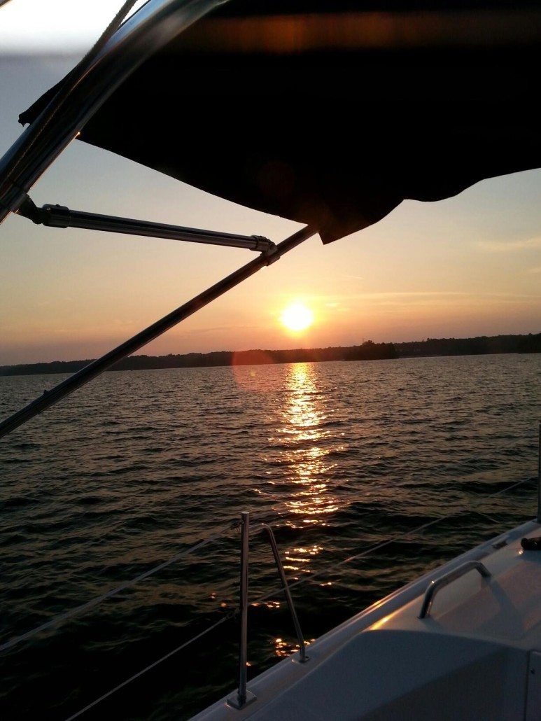 Sunset over water seen from a sailboat. Sun reflects golden light on waves, framed by boat's railing and canopy.