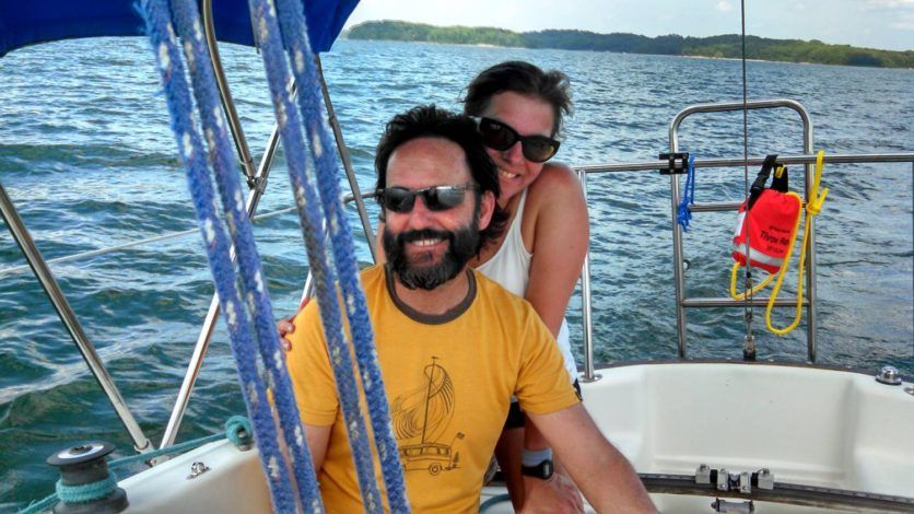 Man and woman on a sailboat, smiling. Man at the helm wearing sunglasses, woman behind him. Blue water and sky.
