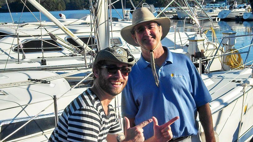 Two men on a sailboat deck, smiling, one wearing a hat, gesturing with hand, other in striped shirt.