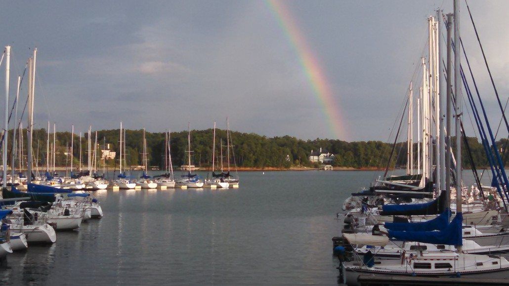 Rainbow over sailboats docked in a harbor, with treed shoreline in the background.