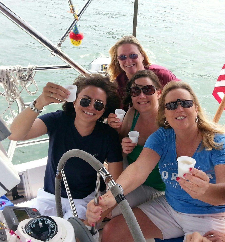 Four women on a sailboat, smiling and holding cups, with one woman at the helm.