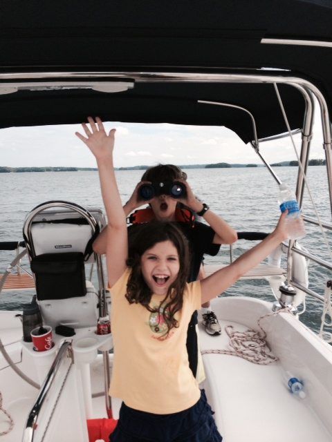 Two children on a sailboat, one with arms raised, other using binoculars, on a lake.