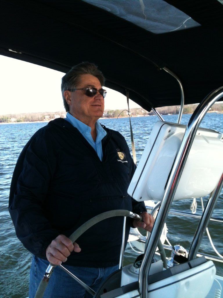 Man steering sailboat on water, wearing sunglasses, blue sweater.