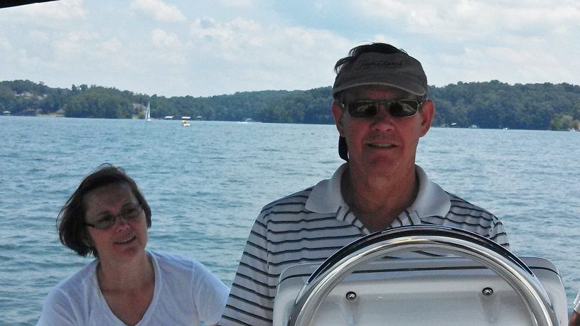 Man steering boat on lake; woman next to him, trees in background.
