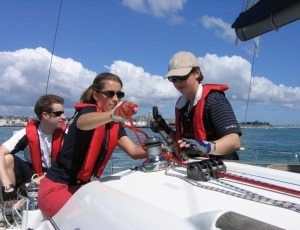 Three people on sailboat, wearing life vests, adjusting ropes. Bright blue sky and water.