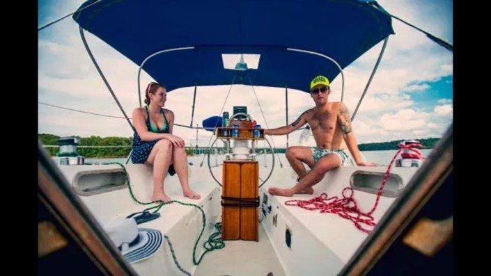 Two people sit on a sailboat. Man steers, woman sits beside him. Blue canopy and sky.