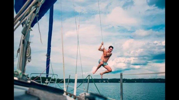 Man swinging on ropes from a sailboat, smiling, above water. Cloudy blue sky.