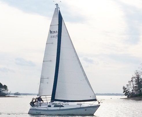 Sailboat on water with white sails; two people visible in the cockpit, trees on the shore in the background.