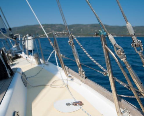 Sailboat deck with rigging against blue water and hills under a clear sky.