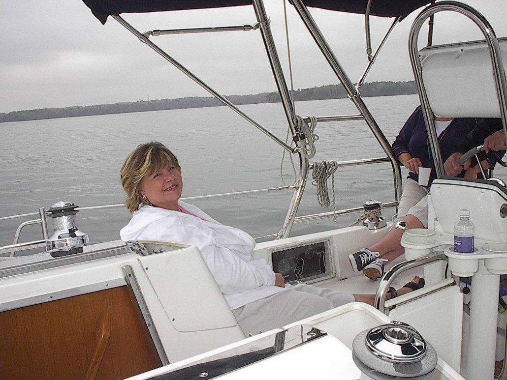 Woman on a sailboat smiles while looking toward the camera. Lake and trees are in the background.
