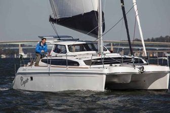Catamaran sailboat with a person on deck, sailing on water near a bridge.
