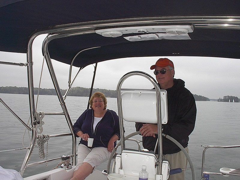 Man steering a sailboat with woman sitting beside him, both smiling on a lake. Overcast, gray sky.