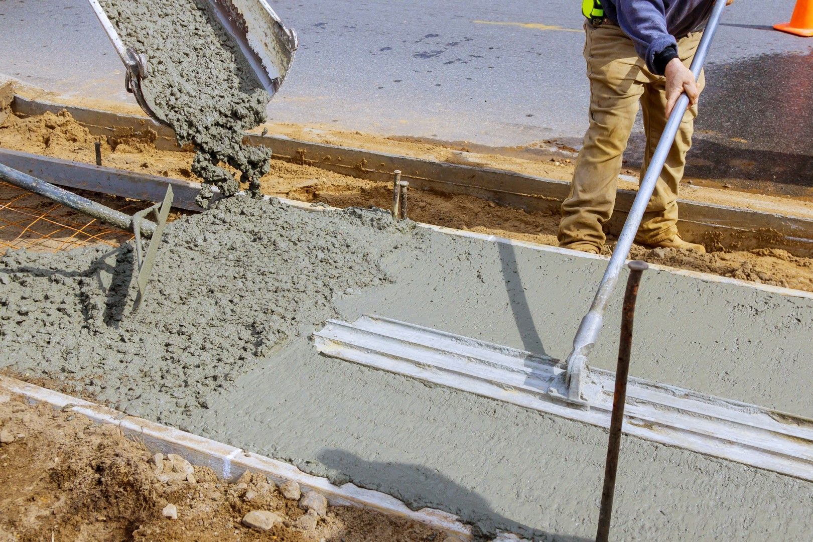 A construction worker pouring and leveling concrete on a sidewalk. The scene includes a street, orange cone, and equipment.