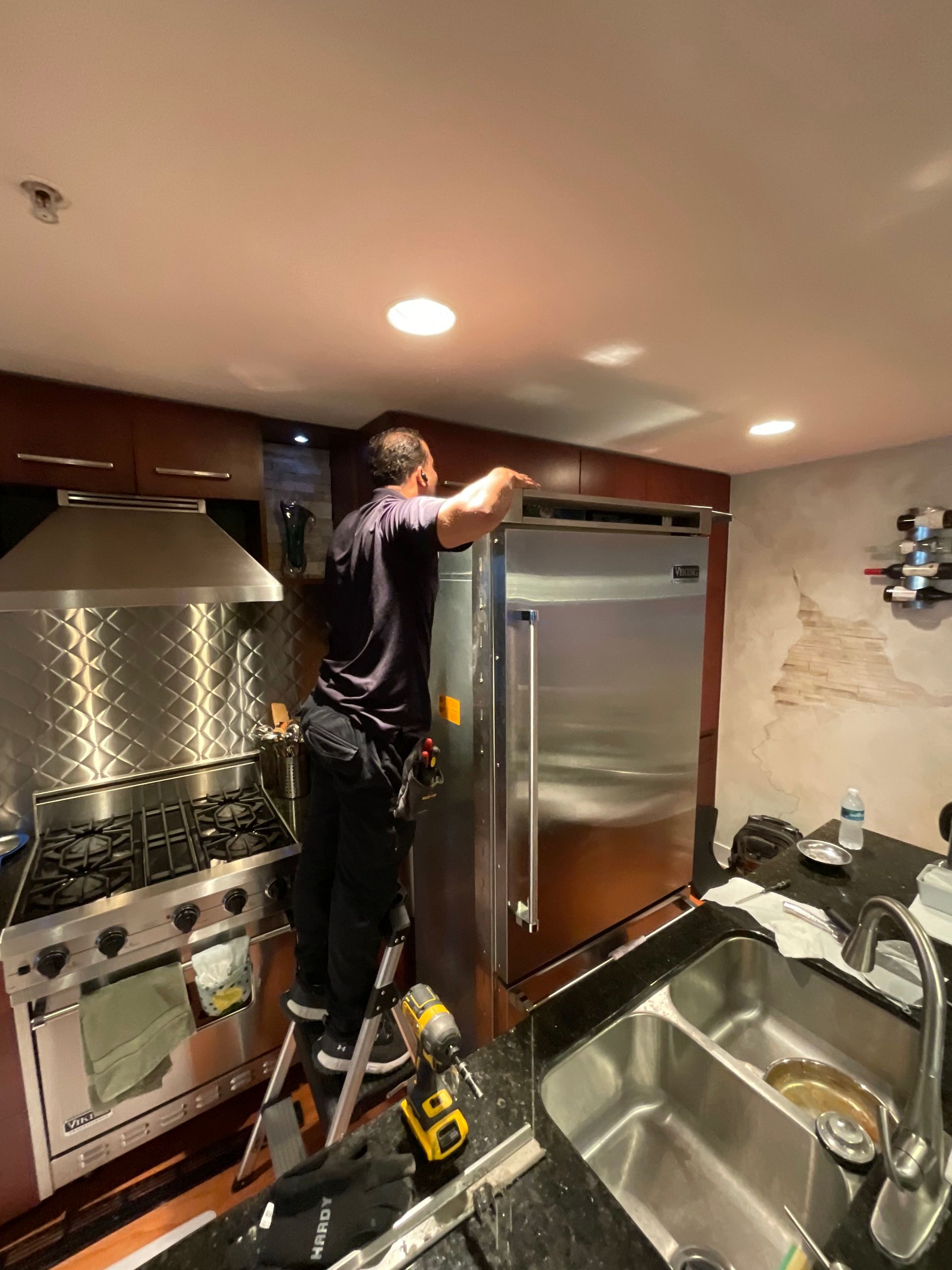A man is standing on a ladder in a kitchen working on a refrigerator.