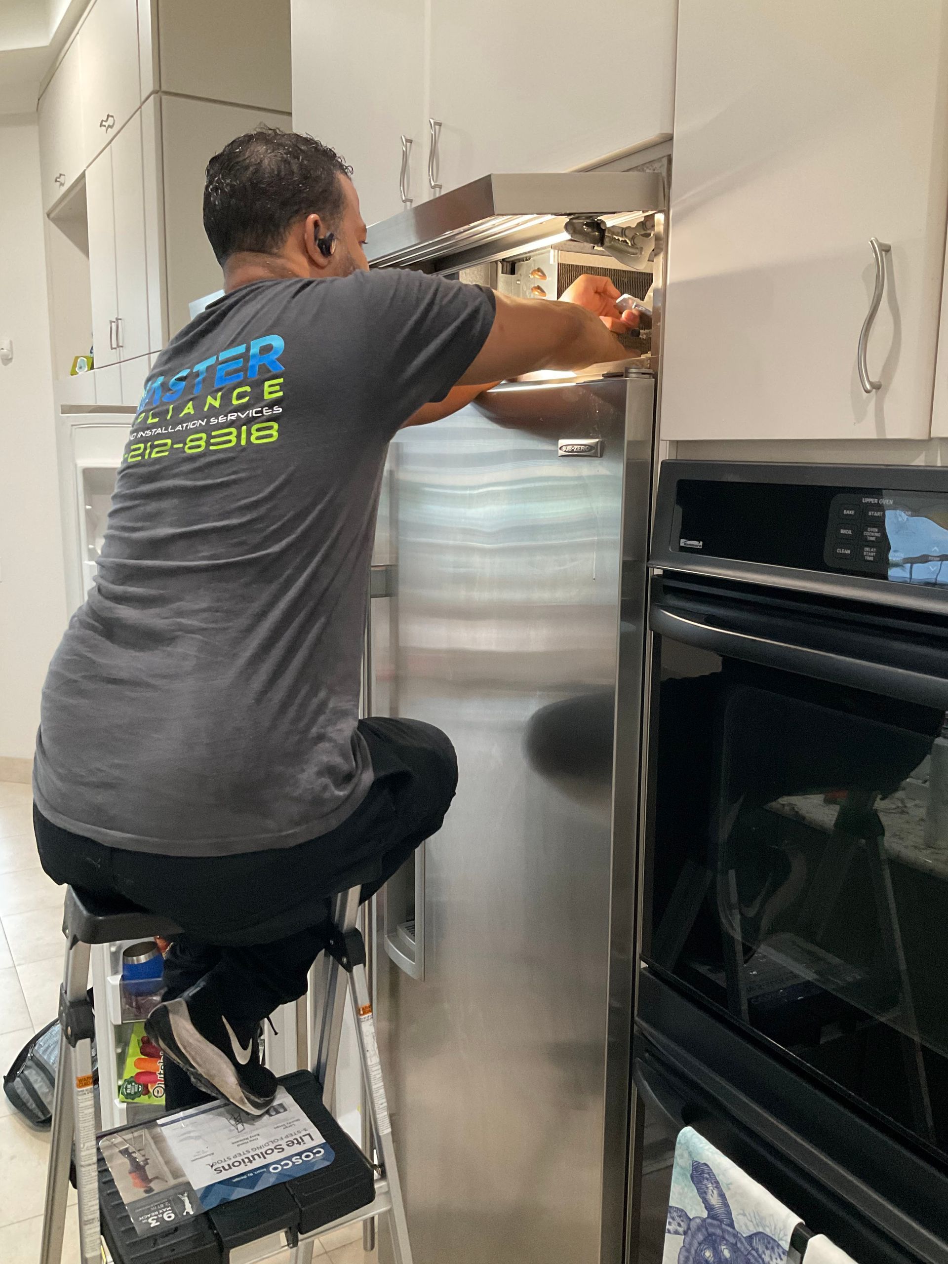 A man is kneeling on a stool working on a refrigerator in a kitchen.