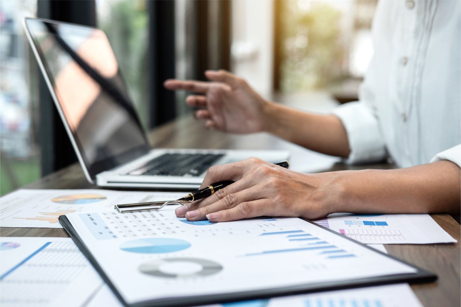 Person pointing at a laptop beside printed charts and documents on a desk