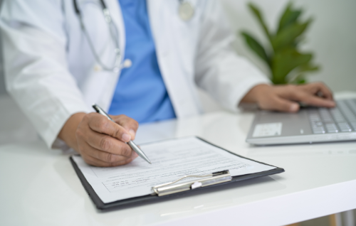 Doctor writing on a clipboard beside a laptop in a clinic