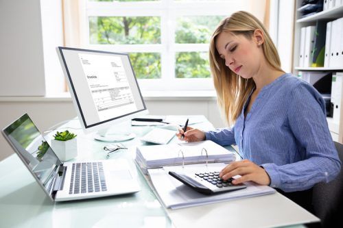 Woman working at a desk with a laptop, desktop monitor, and calculator in a bright office