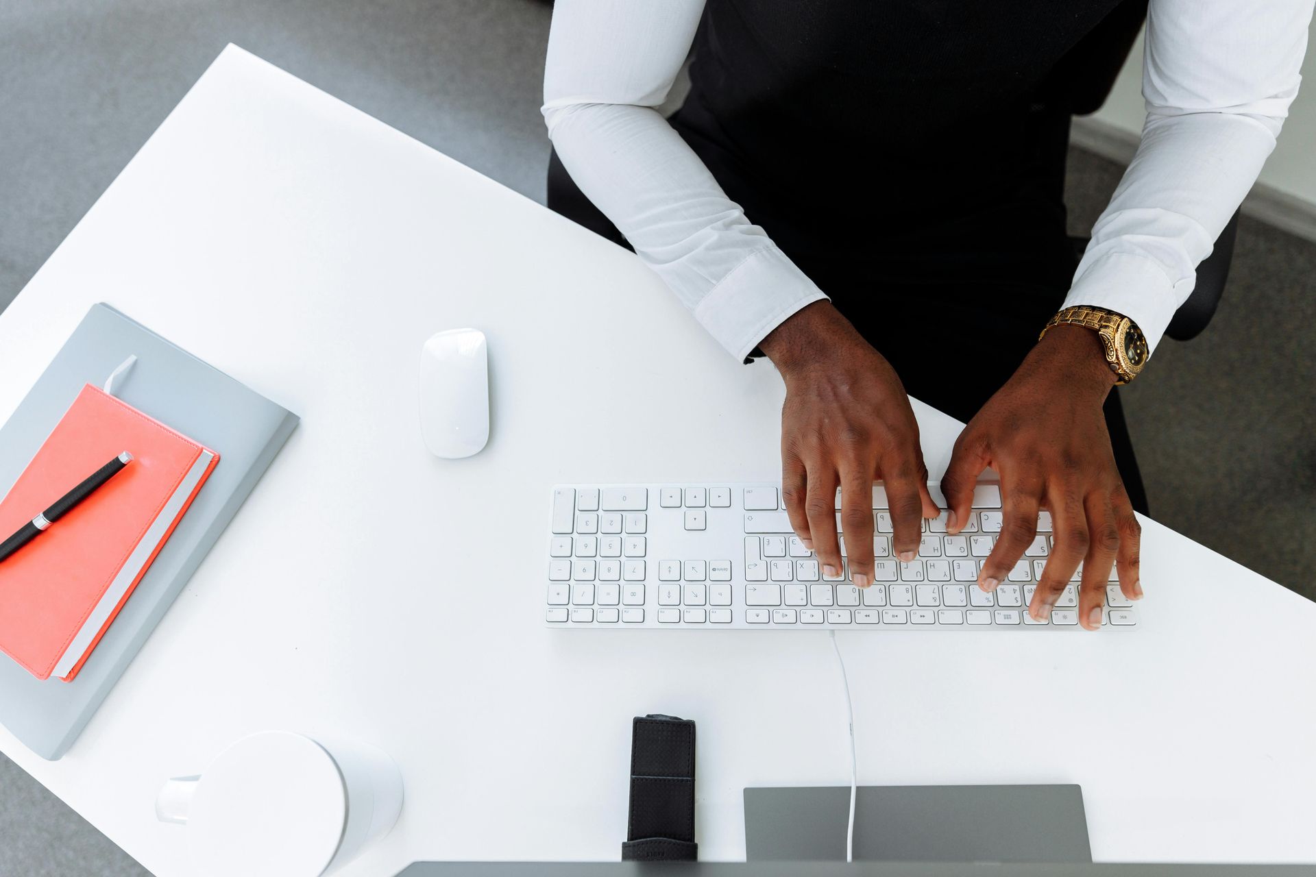 Person typing on a white keyboard at a desk with a laptop, notebook, pen, and coffee mug