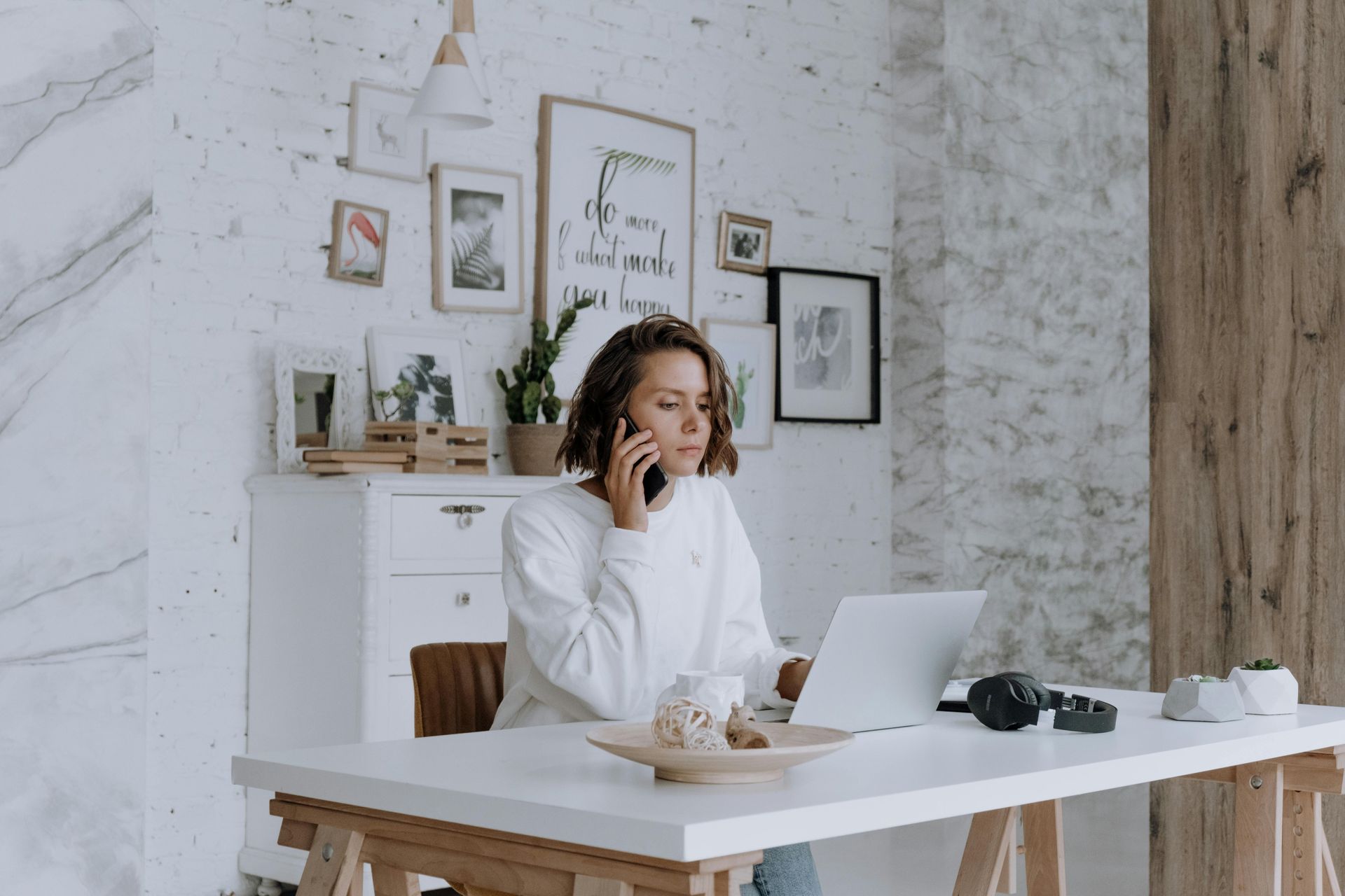 Woman in a white office, smiling on the phone at a desk with a laptop and camera.