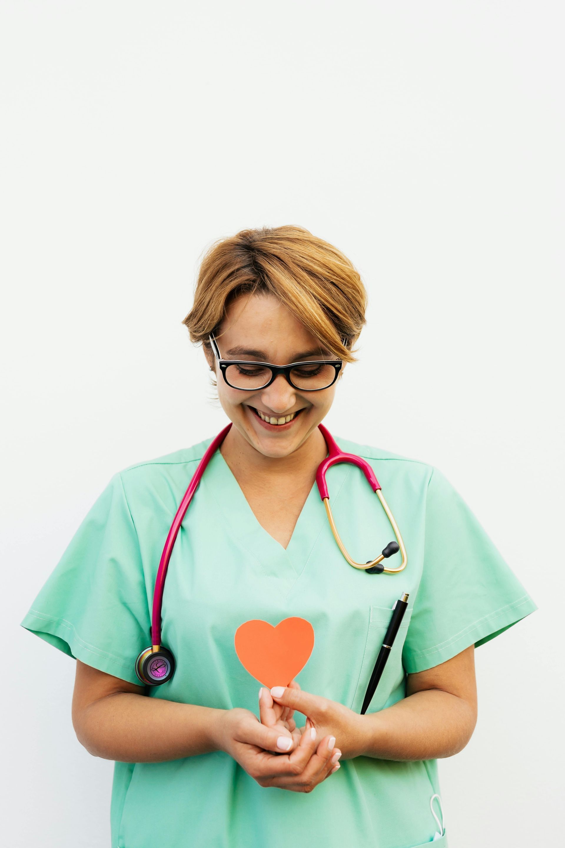 Healthcare worker in green scrubs holding a heart-shaped icon and pen, smiling with stethoscope around neck