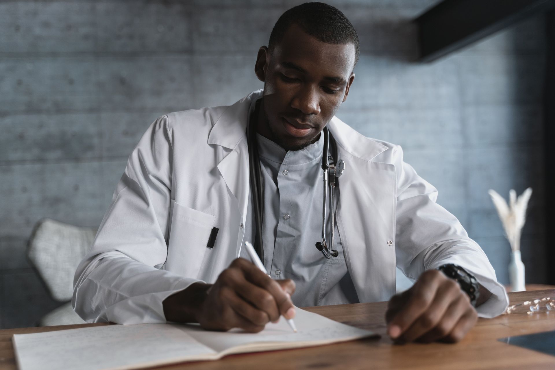 Doctor in a white coat writing notes at a desk with a stethoscope around his neck.