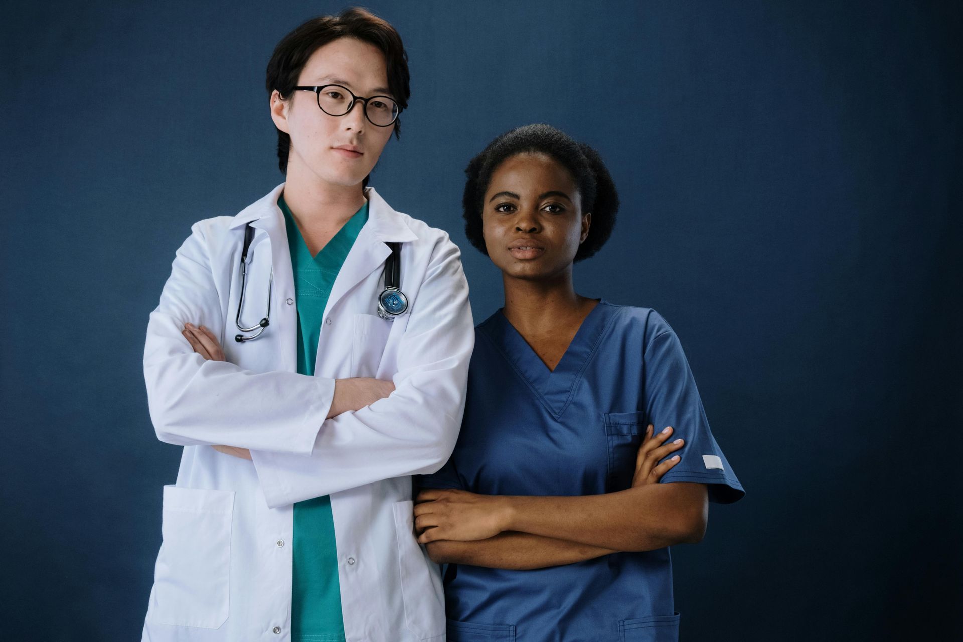 Two healthcare workers in scrubs and lab coat standing with arms crossed against a blue backdrop