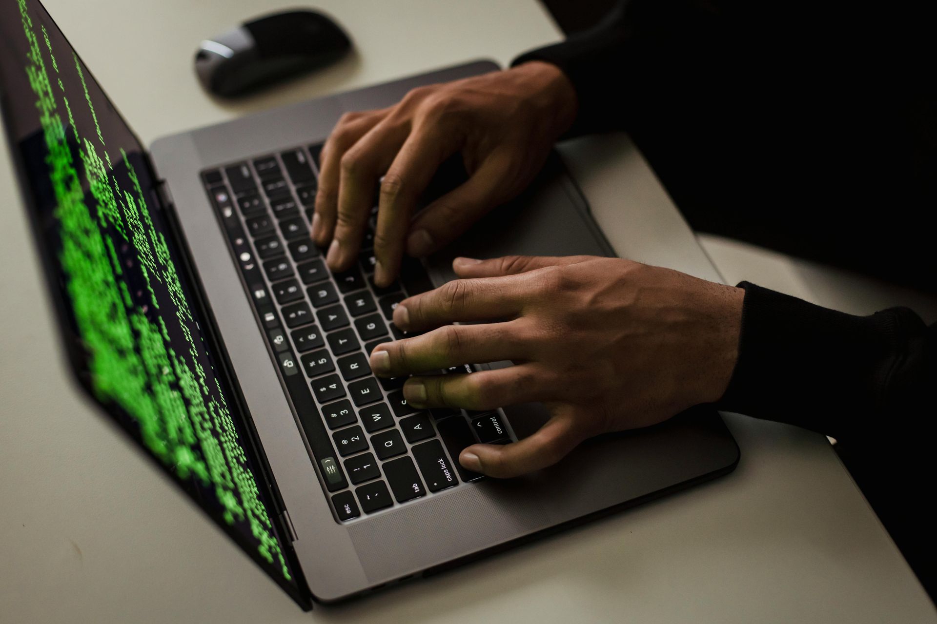 Hands typing on a laptop with green code on the screen, on a desk with a mouse nearby