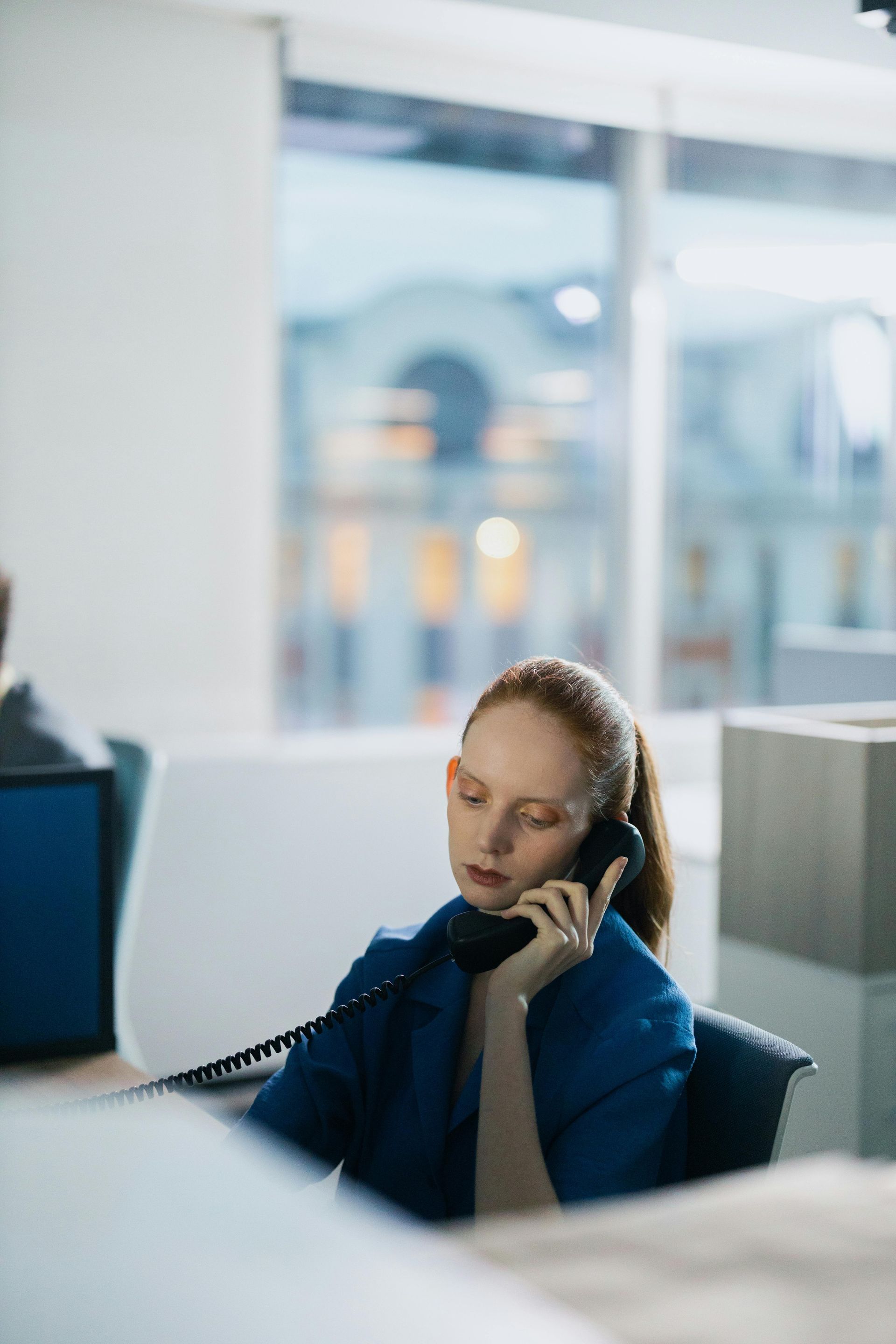Woman in a blue jacket talking on a phone at an office desk with a blurred bright background