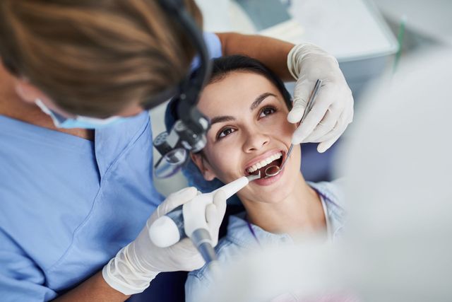 A dentist wearing a mask, face shield, and blue gloves examines a patient in a dental chair under a surgical light.
