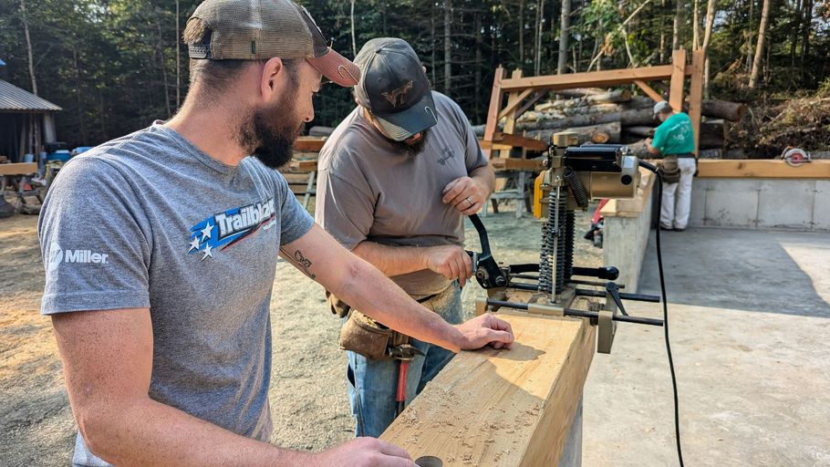Two workers in a workshop measure and cut a wooden beam with a miter saw in bright daylight.