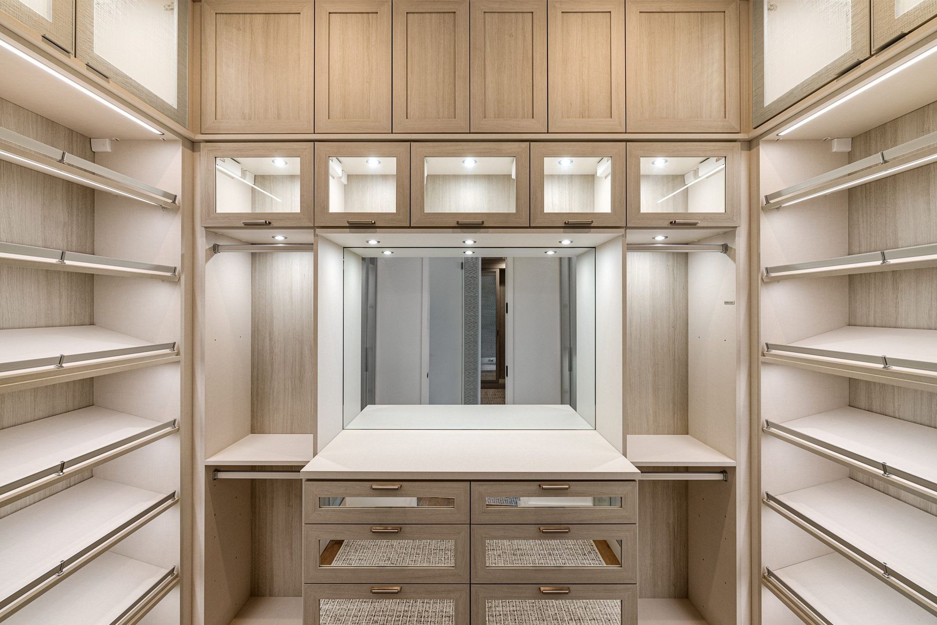 Bright walk-in closet with built-in shelves, drawers, and a central mirrored vanity.