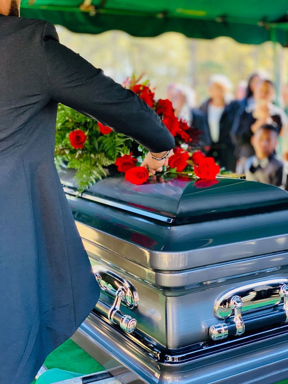 A person places red flowers on a shiny casket at an outdoor funeral.