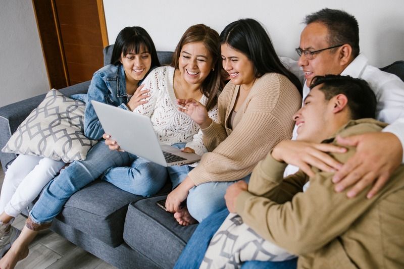 Five people sit on a couch, smiling and looking at a laptop together in a living room.