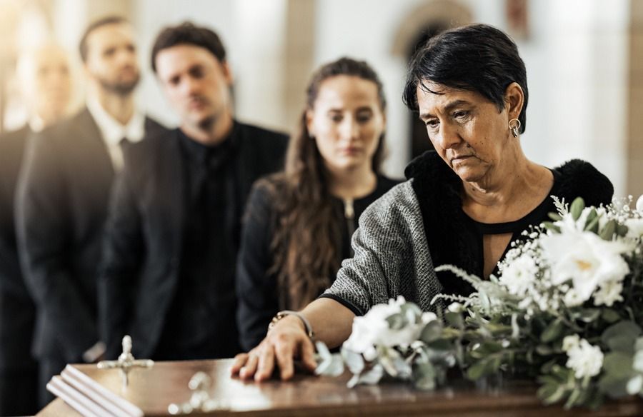 Mourners gathered at a coffin, with a woman touching white flowers in a solemn funeral scene