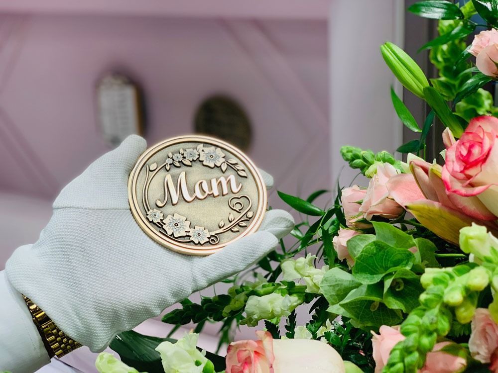 Gloved hand holding a round “Mum” medallion beside pink flowers and greenery