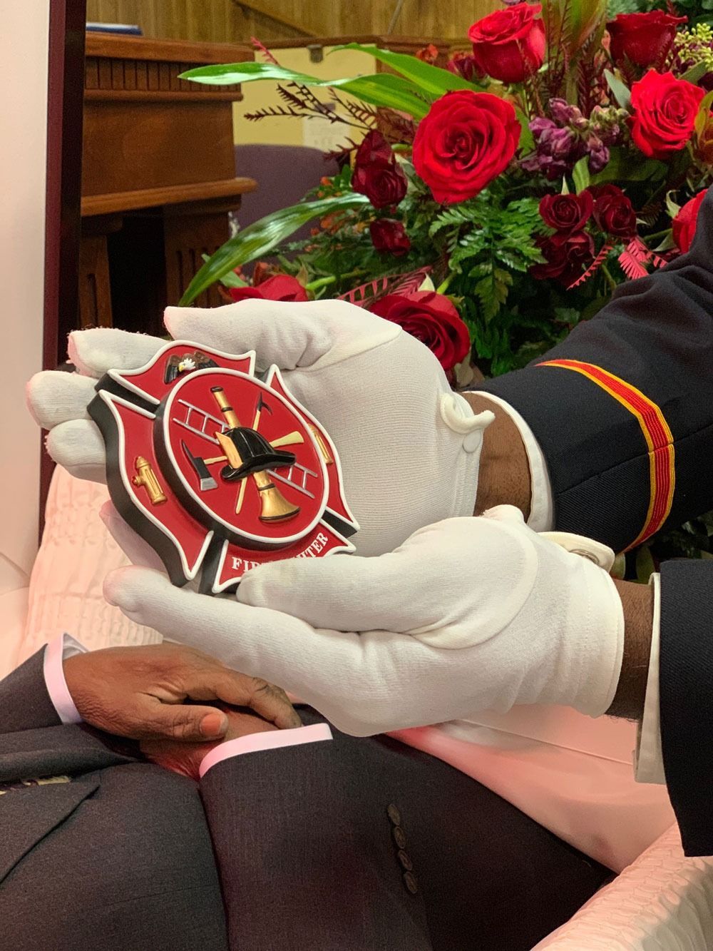 Gloved hands present a red military-style medal with a star emblem beside red roses and a seated person's lap