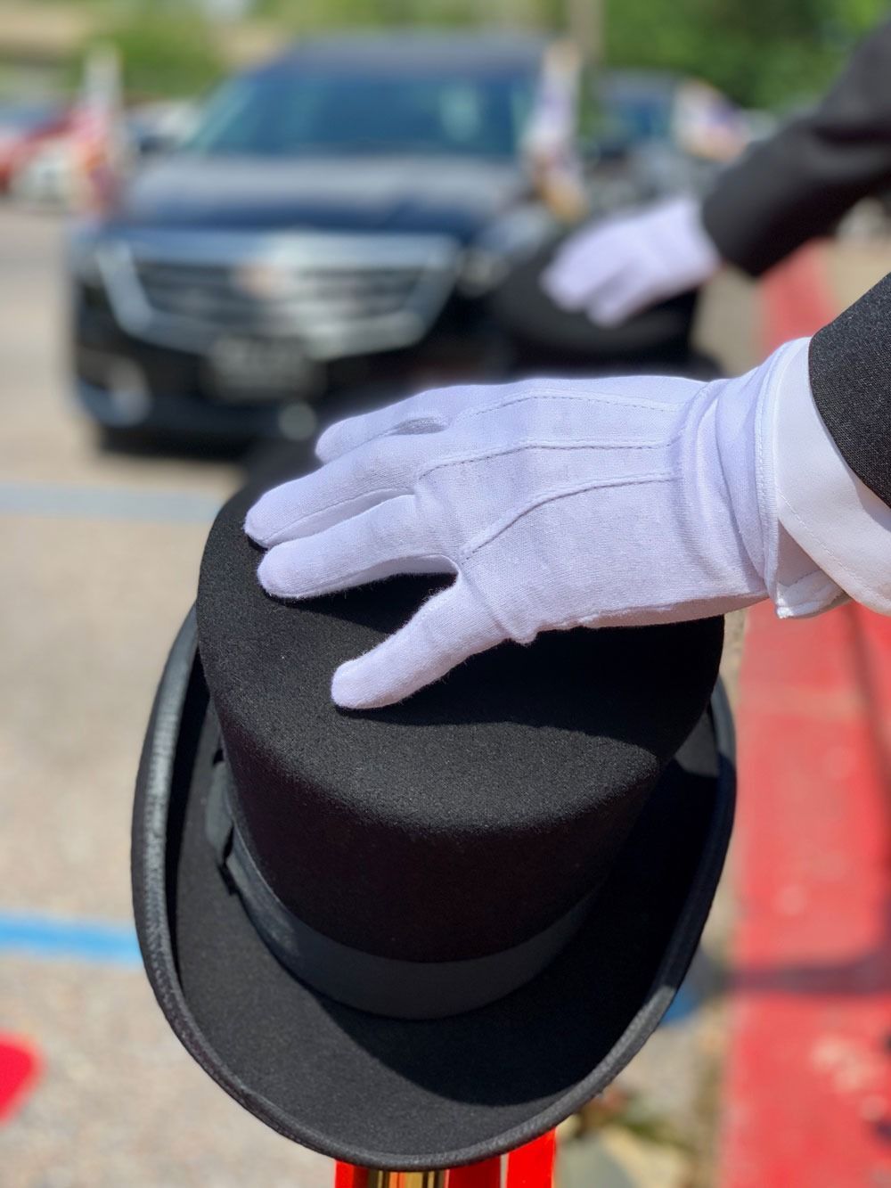 White-gloved hand holding a black top hat in front of a car outdoors