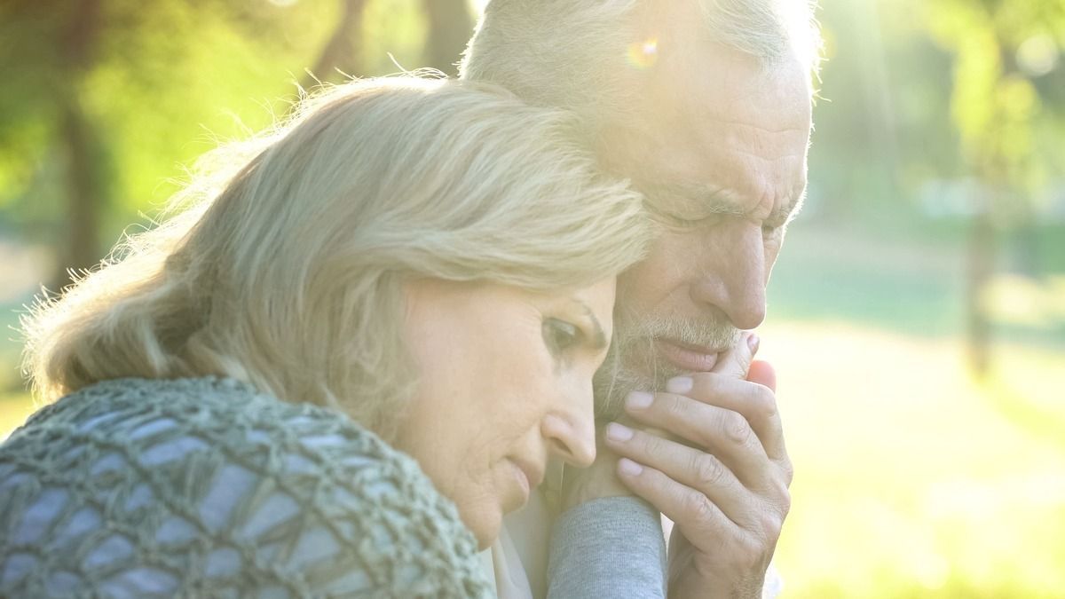Two people embrace outdoors in warm sunlight, with one resting her head on the other’s shoulder.